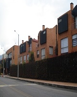 Fotografía de izquierda a derecha vía vehicular seguía un andén peatonal, posteriormente una cerca viva que rodea un conjunto residencial de casas. 