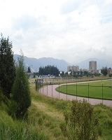 Imagen de zona verde y en la parte intermedia derecha una pista de deporte con una malla de seguridad, al horizonte de la foto se ven conjuntos de vivienda. 